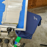 A Shop Shirt | Home Base by Ballad of the Bird Dog in navy rests on a screen printing press, featuring a white dog graphic; similar shirts and printing gear surround the scene.