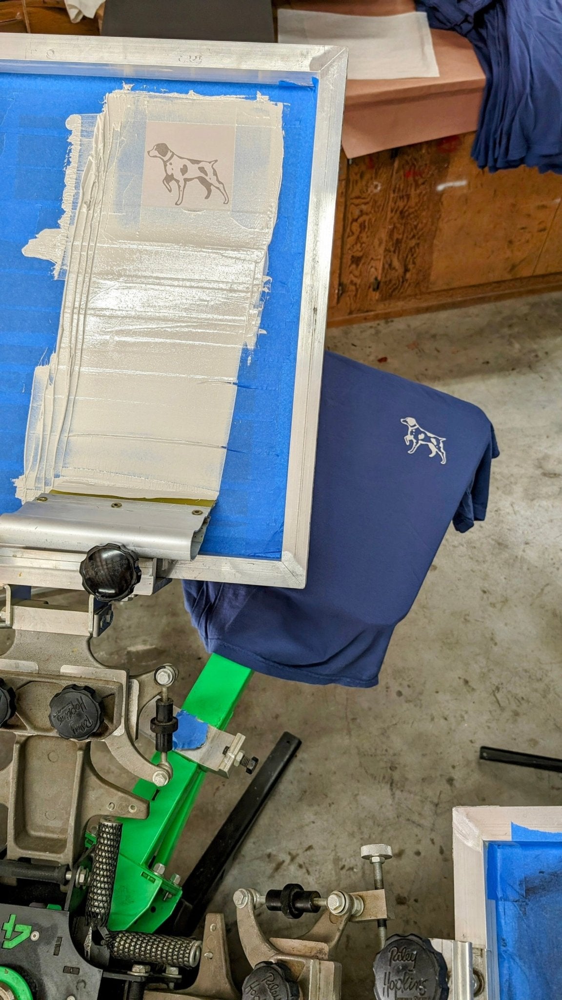 A Shop Shirt | Home Base by Ballad of the Bird Dog in navy rests on a screen printing press, featuring a white dog graphic; similar shirts and printing gear surround the scene.