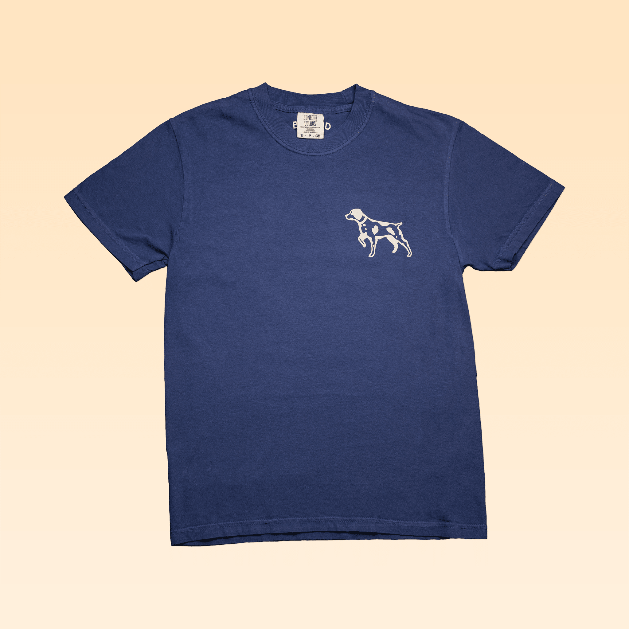 The Shop Shirt | Home Base by Ballad of the Bird Dog is a navy Comfort Colors tee, featuring a small white dog graphic on the left chest and shown on a beige background.