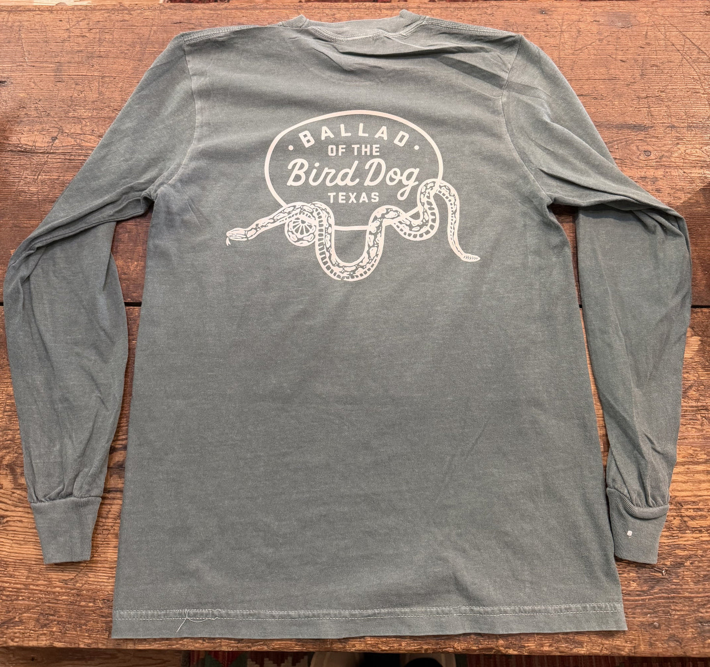 The Ballad of the Bird Dog Shop Shirt Long Sleeve | Rattlesnake Oval is a gray shirt featuring "Ballad of the Bird Dog Texas" and a white snake illustration on the back, displayed on a wooden surface.