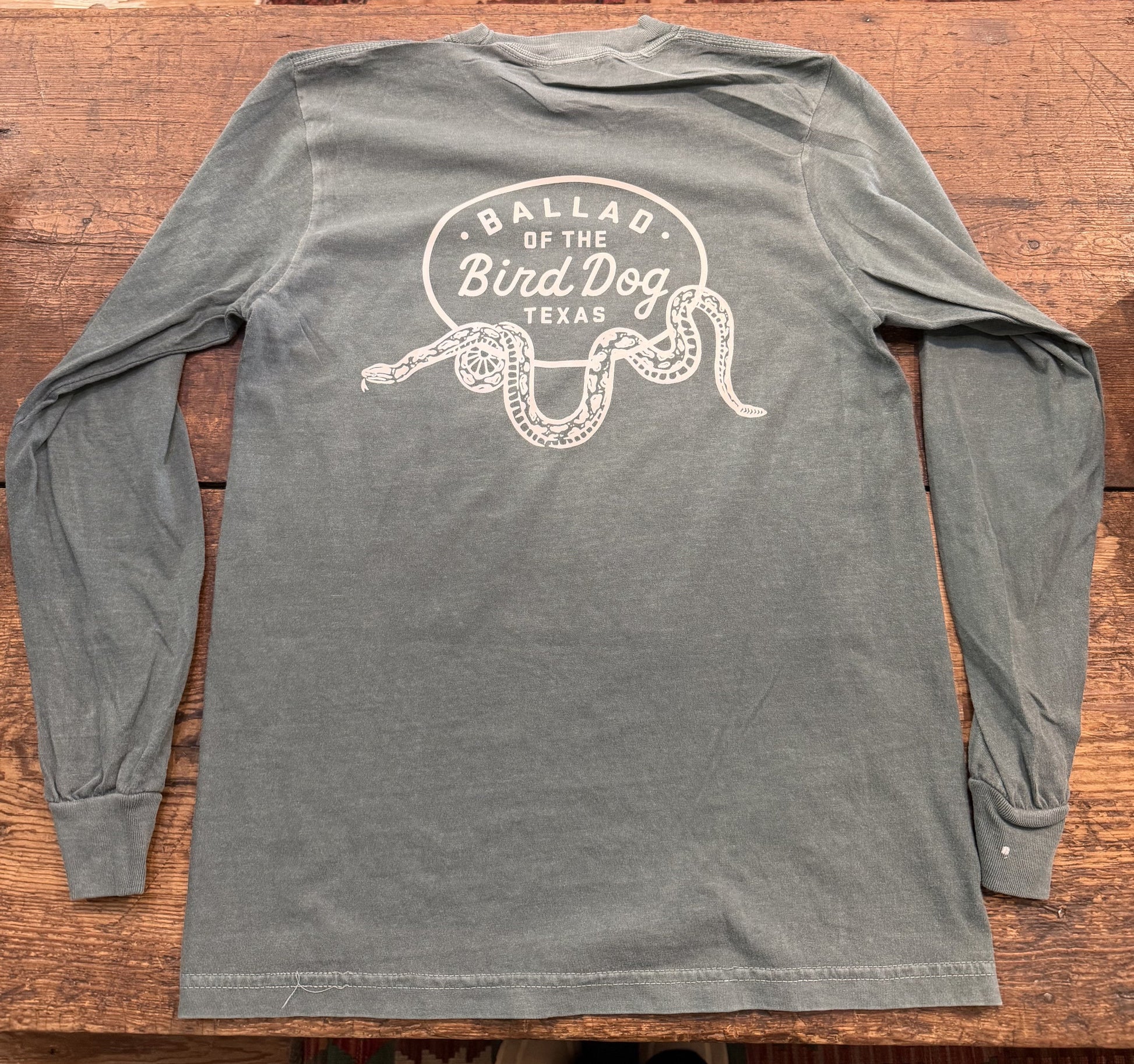 The Ballad of the Bird Dog Shop Shirt Long Sleeve | Rattlesnake Oval is a gray shirt featuring "Ballad of the Bird Dog Texas" and a white snake illustration on the back, displayed on a wooden surface.