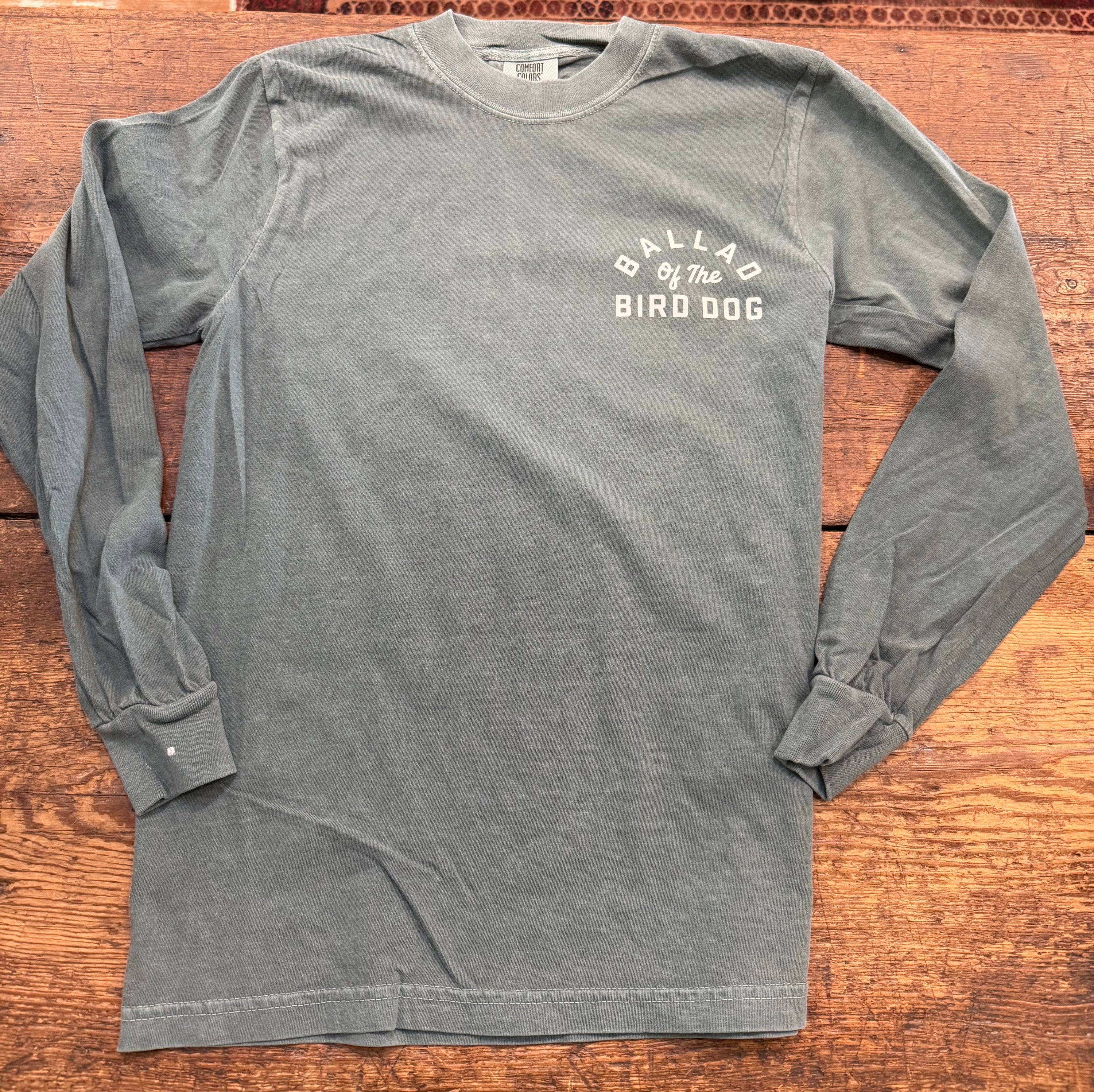 The Ballad of the Bird Dog Shop Shirt Long Sleeve | Rattlesnake Oval features white "Ballad of the Bird Dog" text on the upper left chest and is shown laid flat on a wooden surface.
