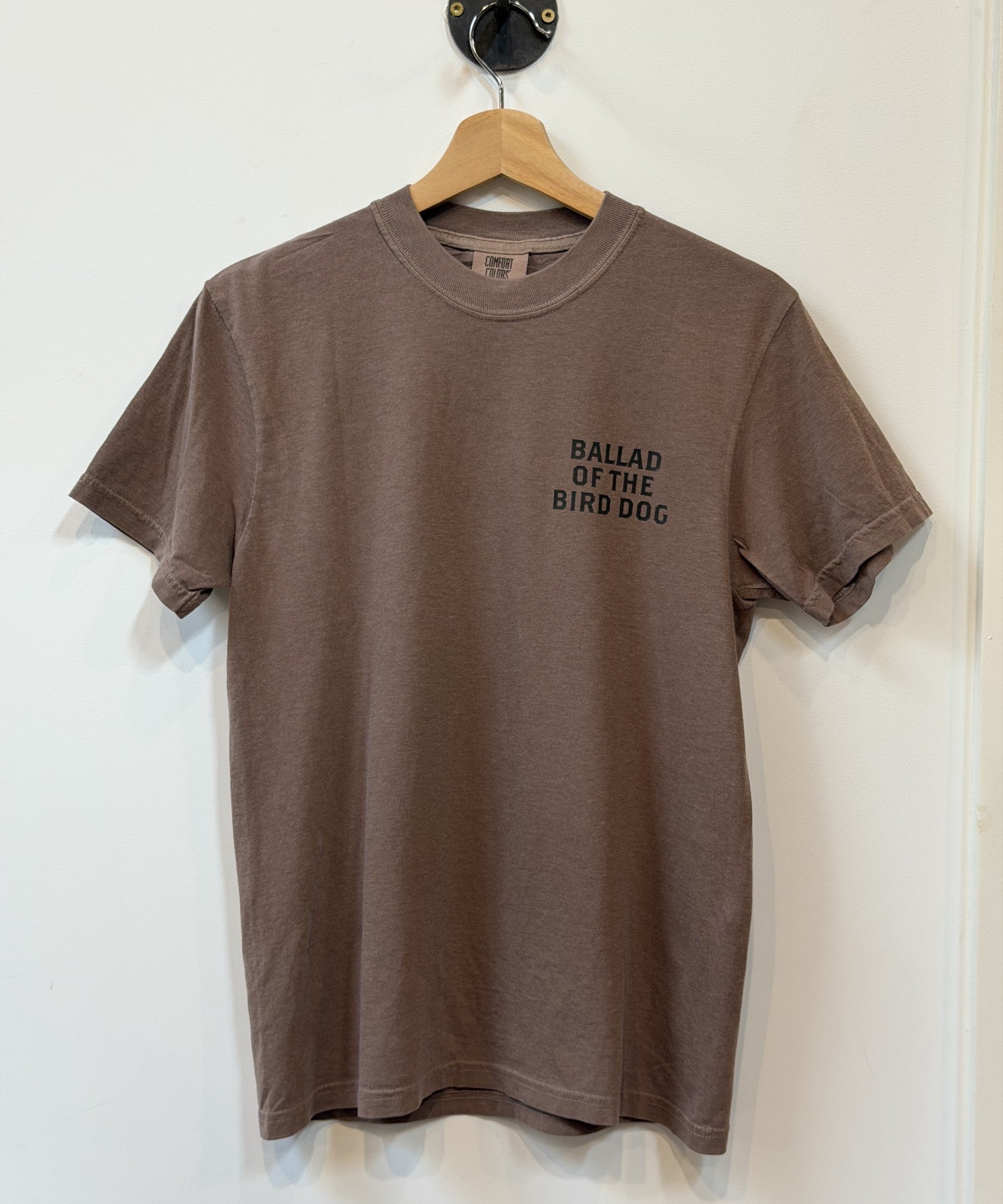 A plain brown Ballad of the Bird Dog "Shop Shirt | Loyal to The Land" t-shirt on a hanger, featuring "BALLAD OF THE BIRD DOG" printed in black on the left chest.