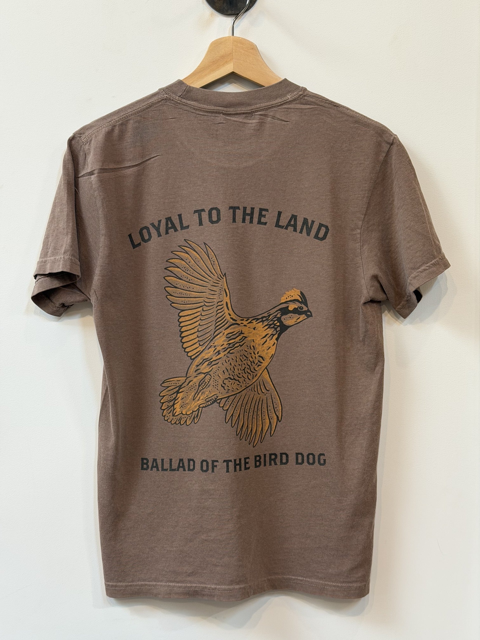 The Shop Shirt by Ballad of the Bird Dog features a brown design with a bird in flight illustration and "LOYAL TO THE LAND" and "BALLAD OF THE BIRD DOG" printed on the back.