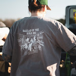 A person in a green cap and a Ballad of the Bird Dog Shop Shirt | Old Fashioned stands outdoors by a table with bottles, highlighting unique Southern Arizona style from Ballad of the Bird Dog.
