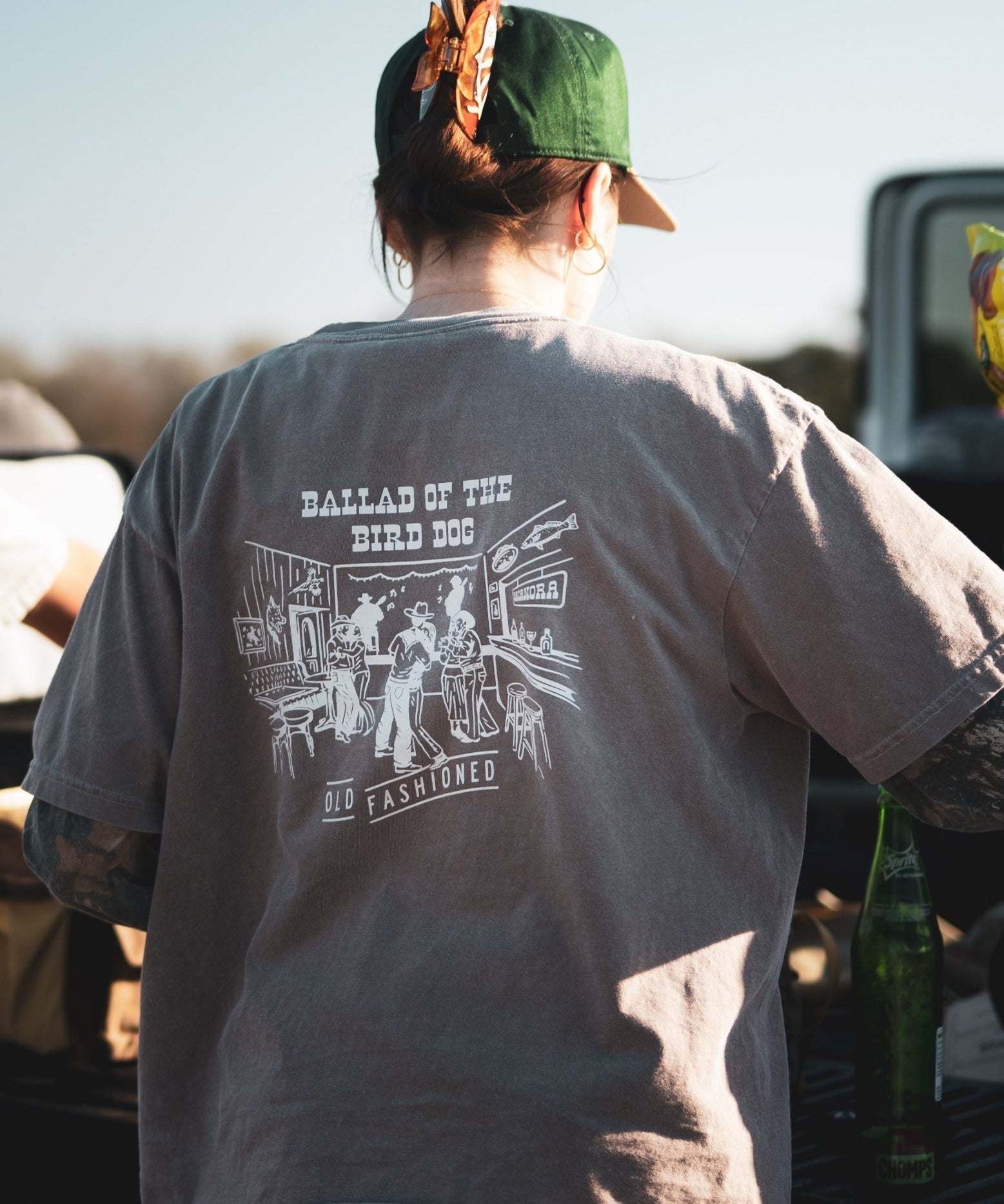 A person in a green cap and a Ballad of the Bird Dog Shop Shirt | Old Fashioned stands outdoors by a table with bottles, highlighting unique Southern Arizona style from Ballad of the Bird Dog.