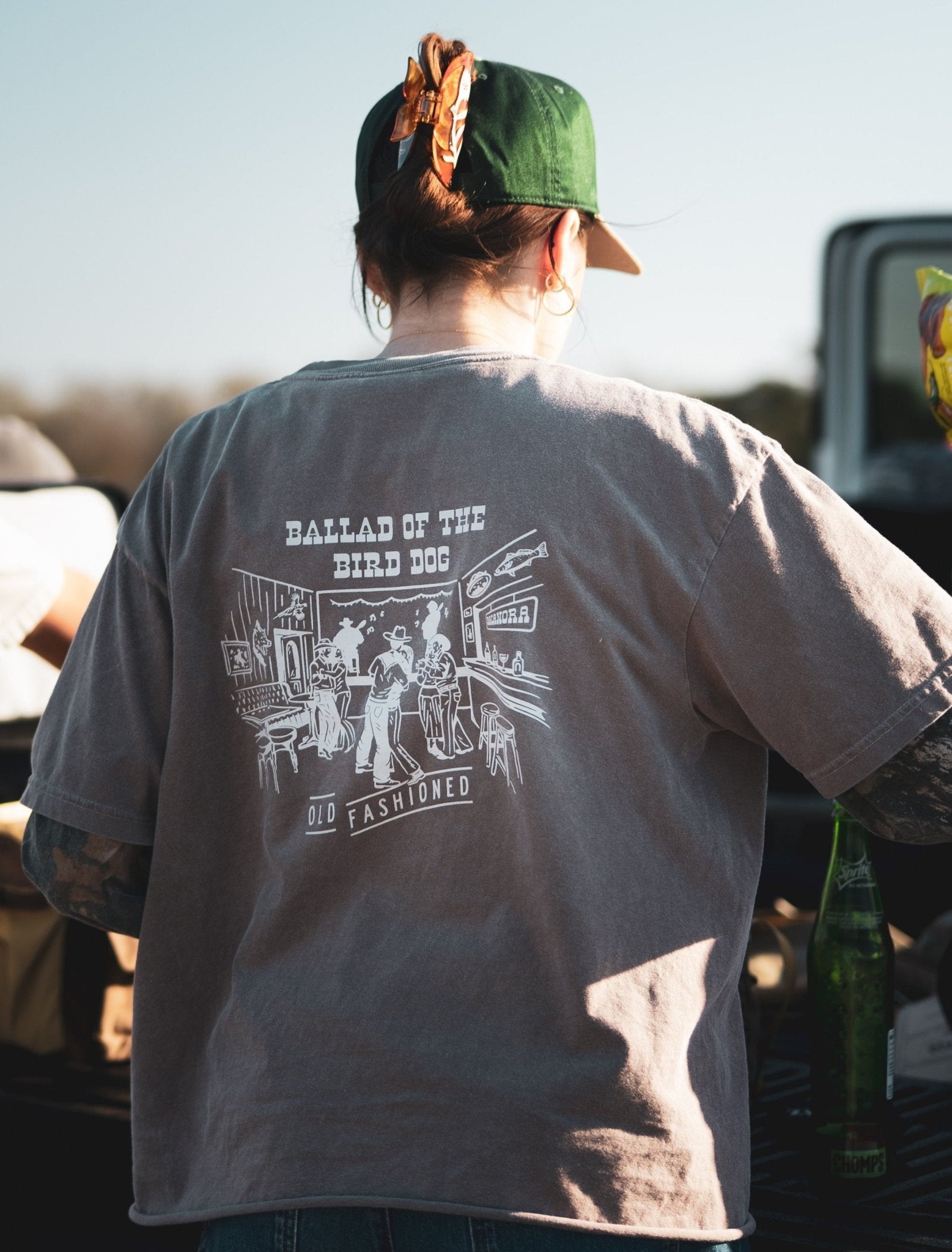 A person in a green cap and a Ballad of the Bird Dog Shop Shirt | Old Fashioned stands outdoors by a table with bottles, highlighting unique Southern Arizona style from Ballad of the Bird Dog.