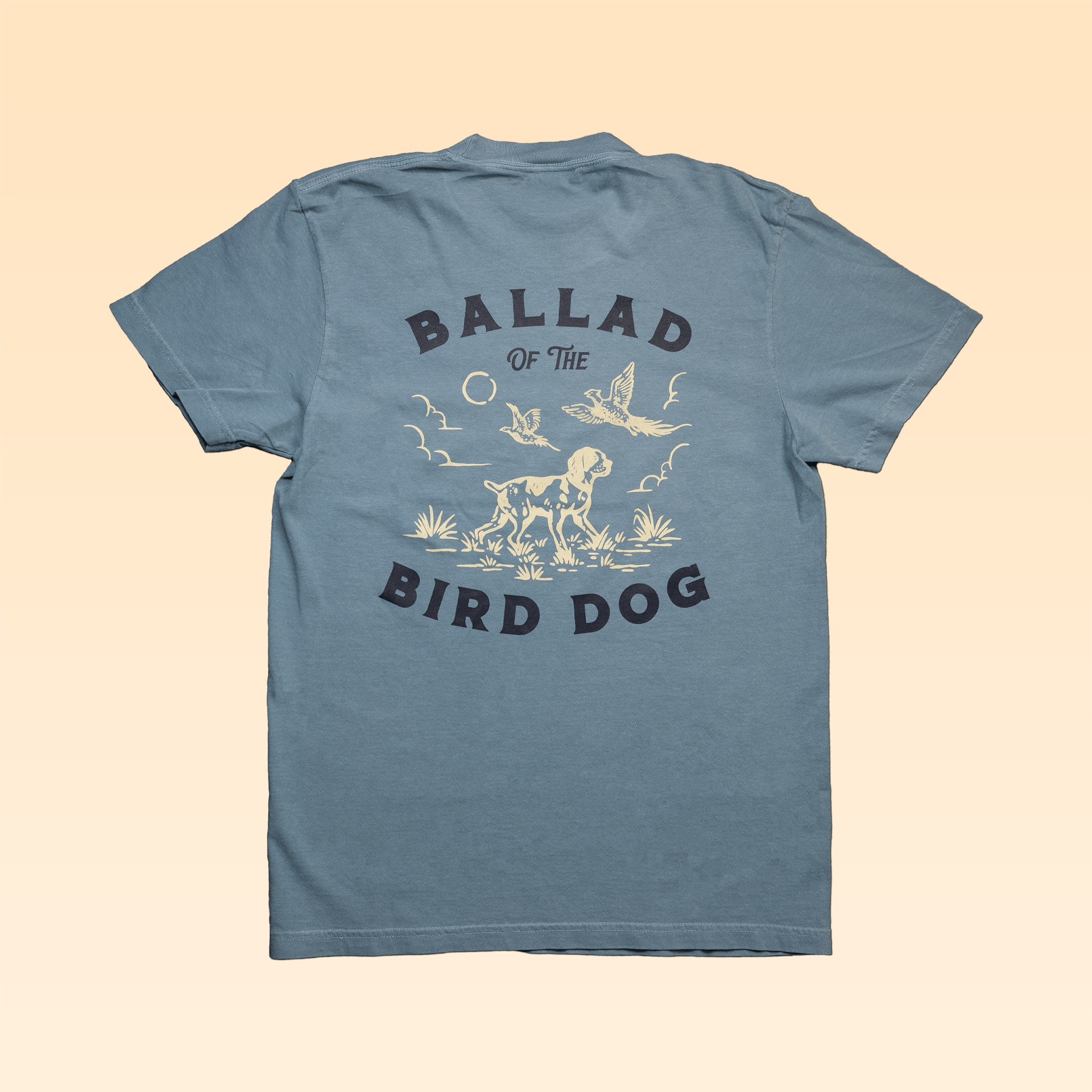 The Ballad of the Bird Dog "Shop Shirt | Pheasant Flush" is a blue vintage-style tee featuring "BALLAD OF THE BIRD DOG" text and a bird dog, birds, sun, and plants graphic on the back.