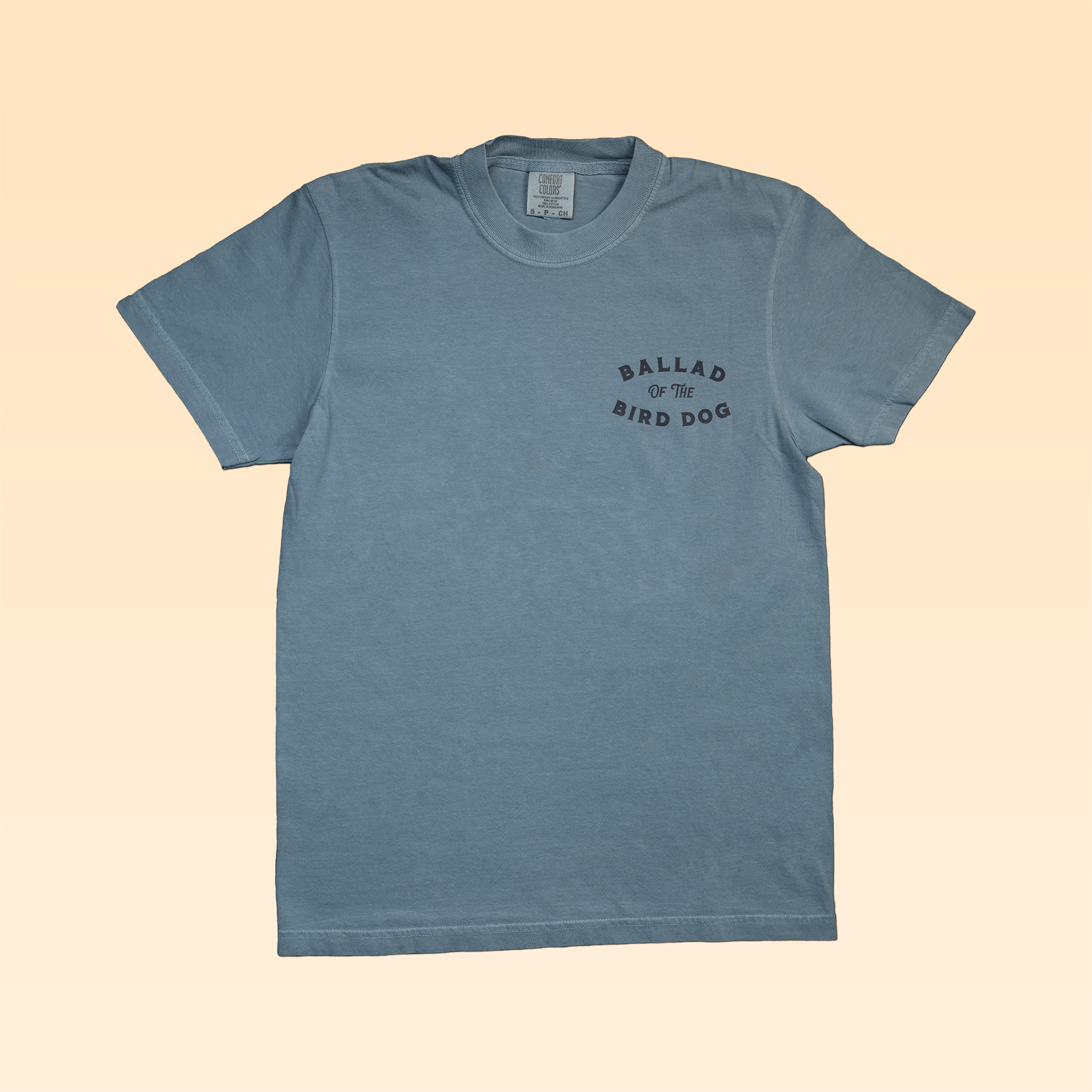 A light blue Shop Shirt | Pheasant Flush by Ballad of the Bird Dog, short-sleeve with "BALLAD OF THE BIRD DOG" in black on the left chest, shown on a beige background.