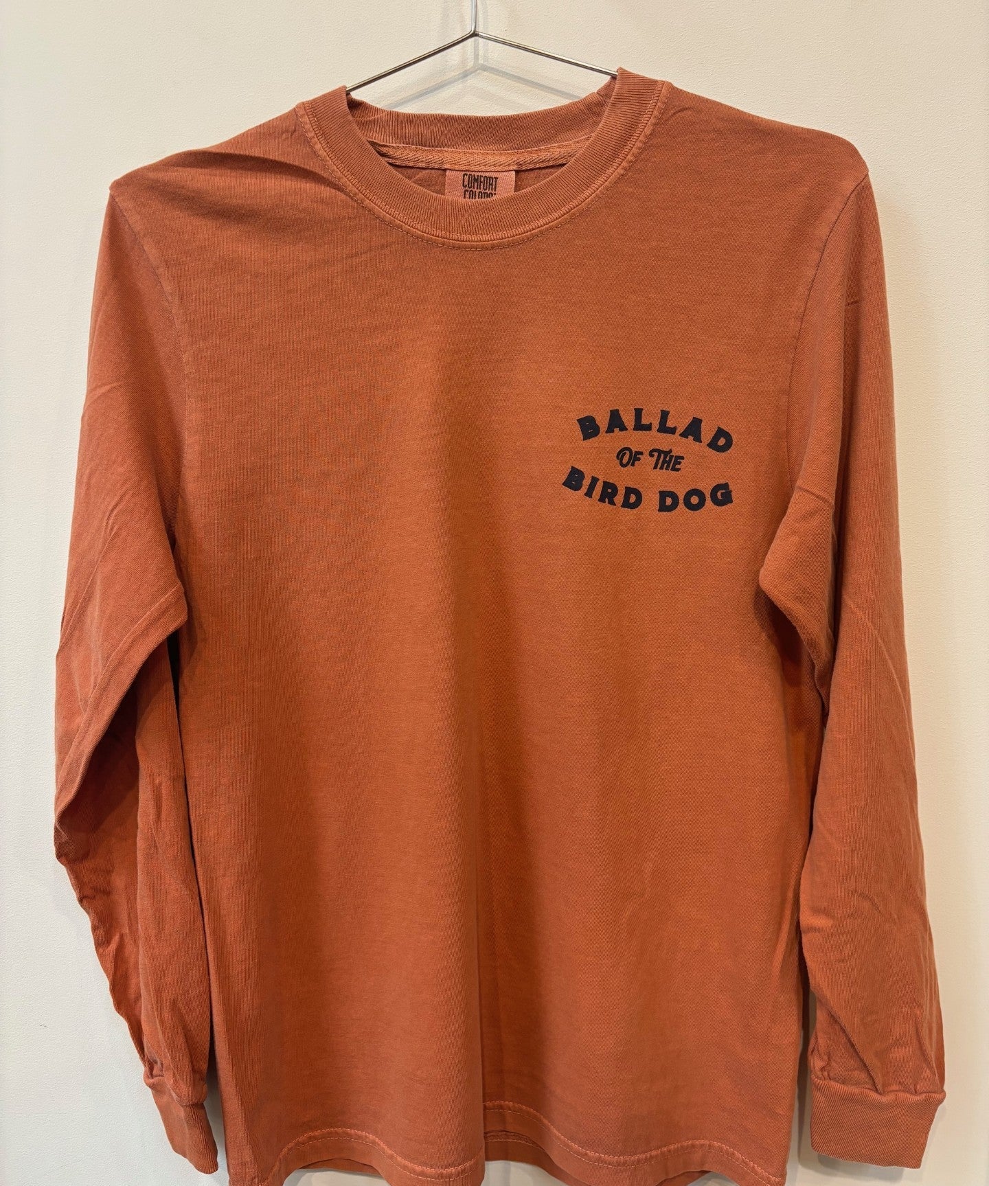 The Shop Shirt | Pheasant Flush by Ballad of the Bird Dog is a rust-colored long-sleeve shirt with "Ballad of the Bird Dog" printed in black on the left chest, shown hanging on a hanger.
