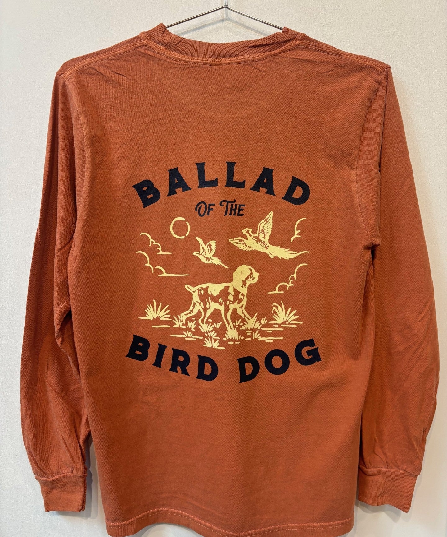 The Shop Shirt | Pheasant Flush by Ballad of the Bird Dog is a rust-colored long-sleeve featuring a dog and flying birds design, with “Ballad of the Bird Dog” printed on the back.