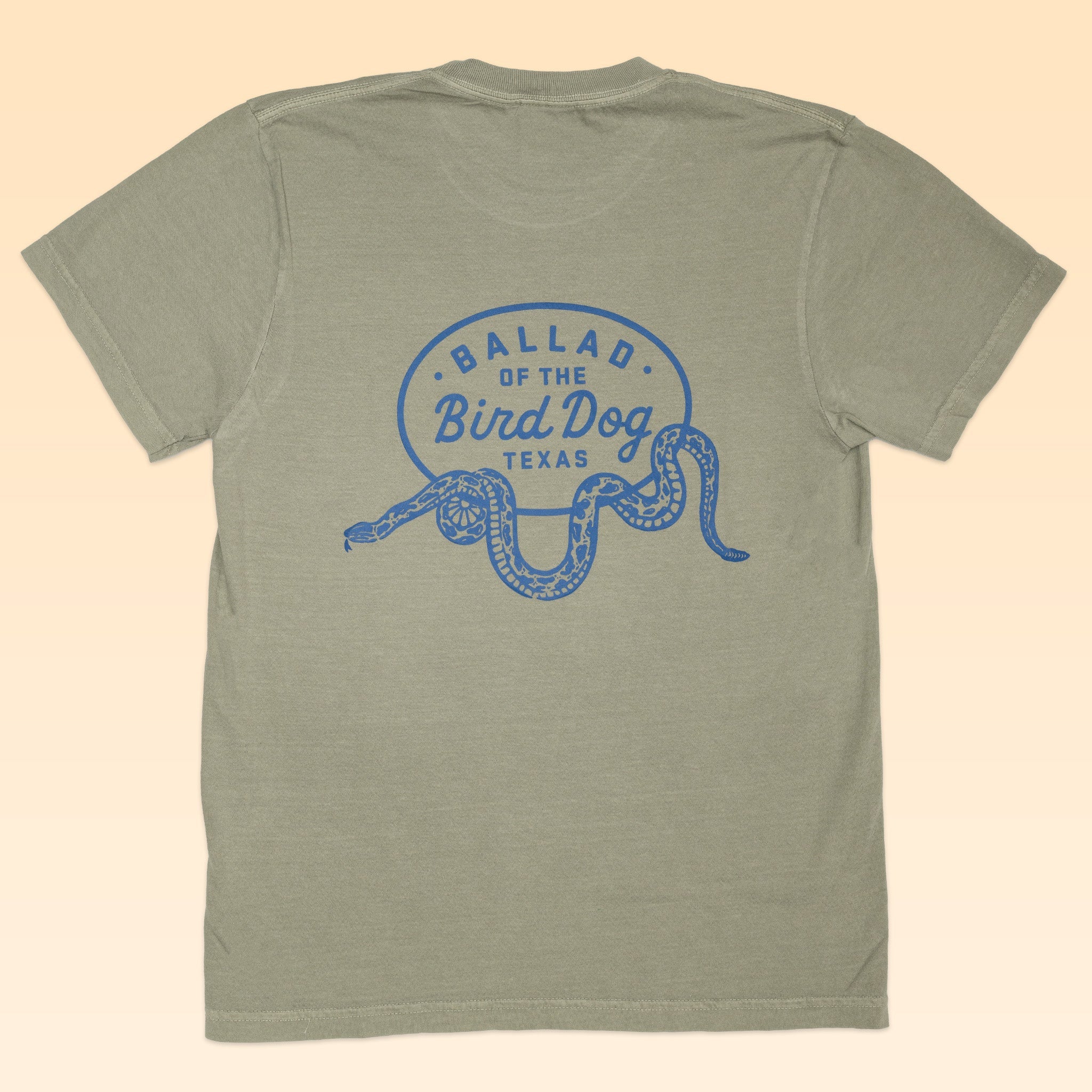 A light olive green Shop Shirt by Ballad of the Bird Dog featuring a blue rattlesnake oval logo and "Ballad of the Bird Dog Texas" on the back—ideal for fans of unique, snake-themed apparel.