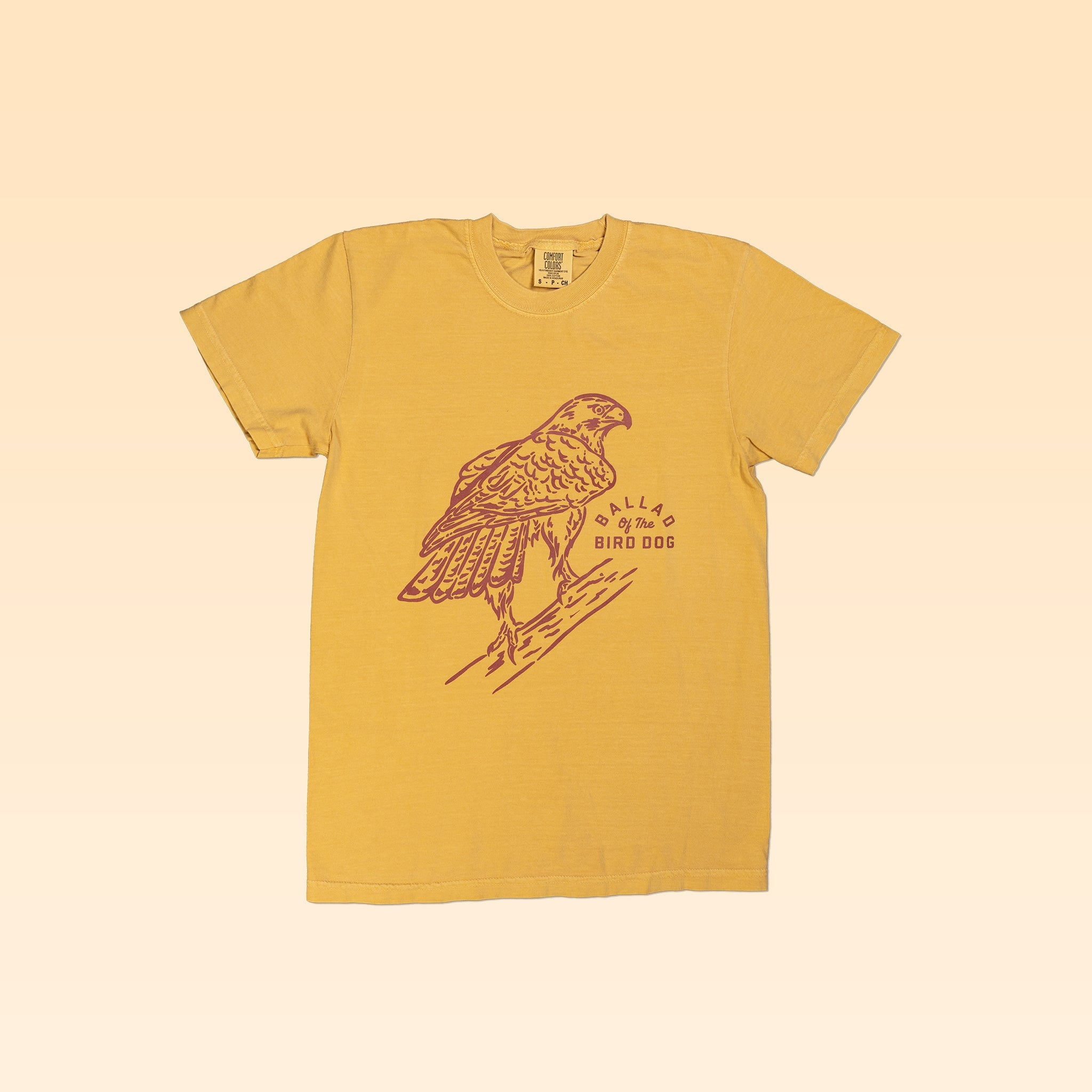 The Shop Shirt from Ballad of the Bird Dog’s Desert Scenes Collection is a yellow tee featuring a red-tailed hawk illustration and "DILLAS BY THE BIRD DOG" in brown, crafted with soft USA cotton.