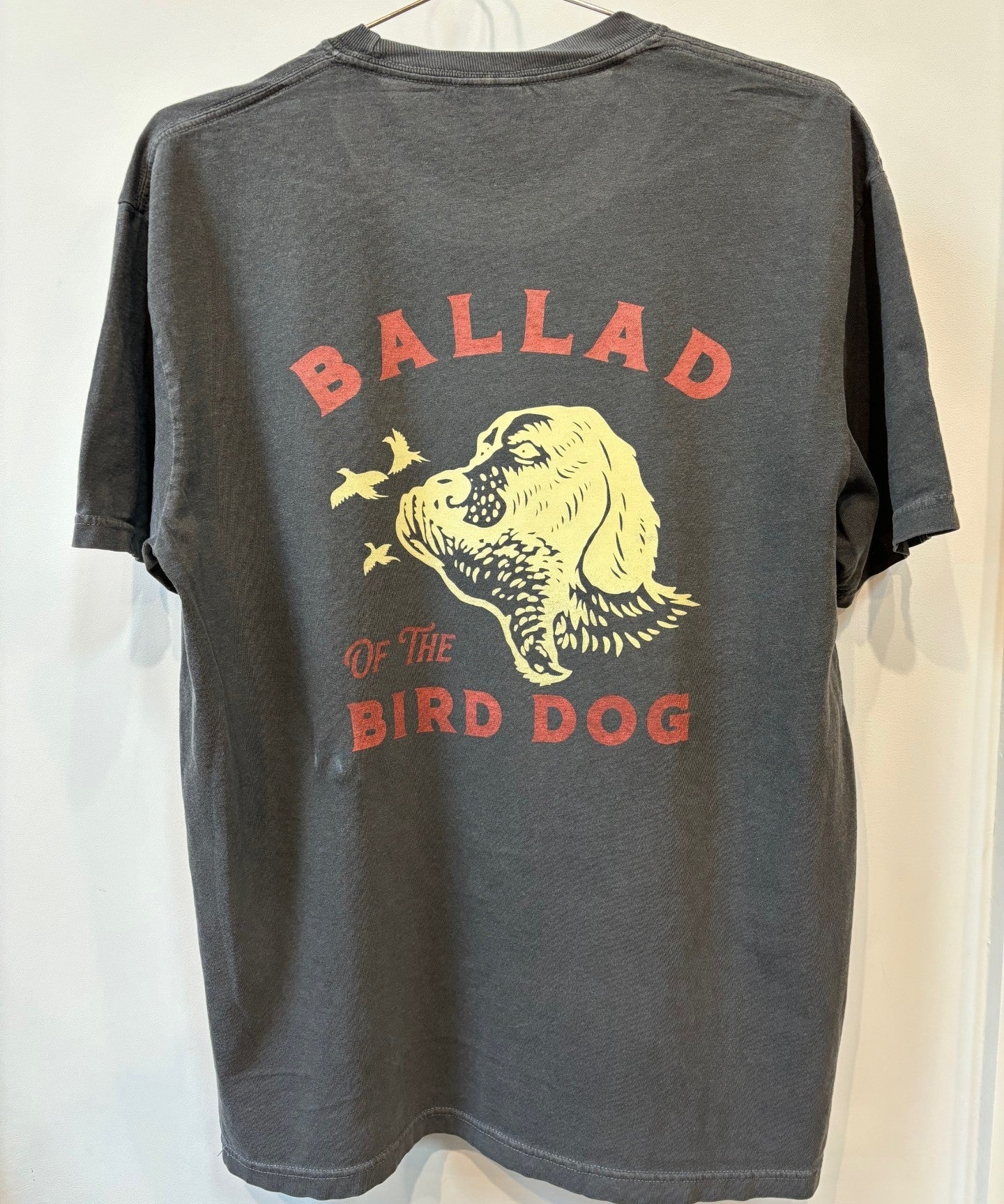 The Shop Shirt | Retriever by Ballad of the Bird Dog is a dark gray tee featuring a yellow dog's head with flying birds and "Ballad of the Bird Dog" printed in red on the back.