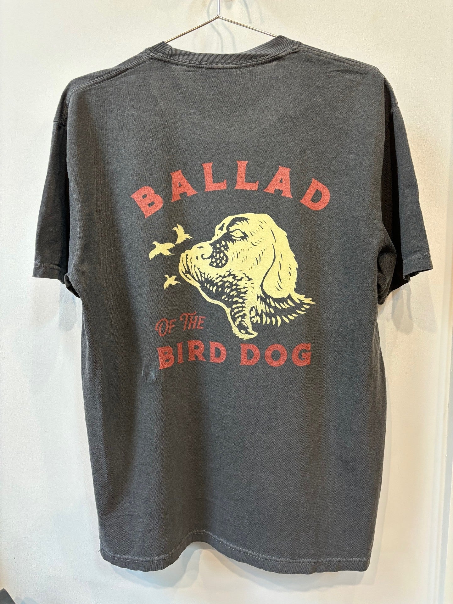 The Shop Shirt | Retriever by Ballad of the Bird Dog is a dark gray tee featuring a yellow dog's head with flying birds and "Ballad of the Bird Dog" printed in red on the back.