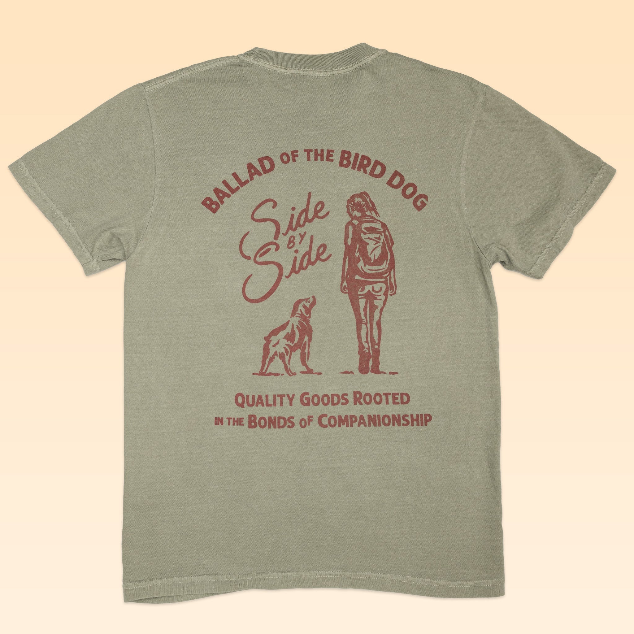 The Shop Shirt | Side By Side by Ballad of the Bird Dog is a khaki shirt with maroon print featuring a dog and person side by side, and the text: “BALLAD OF THE BIRD DOG, Side by Side, Quality Goods Rooted in the Bonds of Companionship.”.