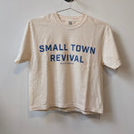 A beige Ballad of the Bird Dog Shop Shirt | Small Town Revival Crop hangs on a hook, featuring bold blue "SMALL TOWN REVIVAL" text—ideal for showing your small town pride.