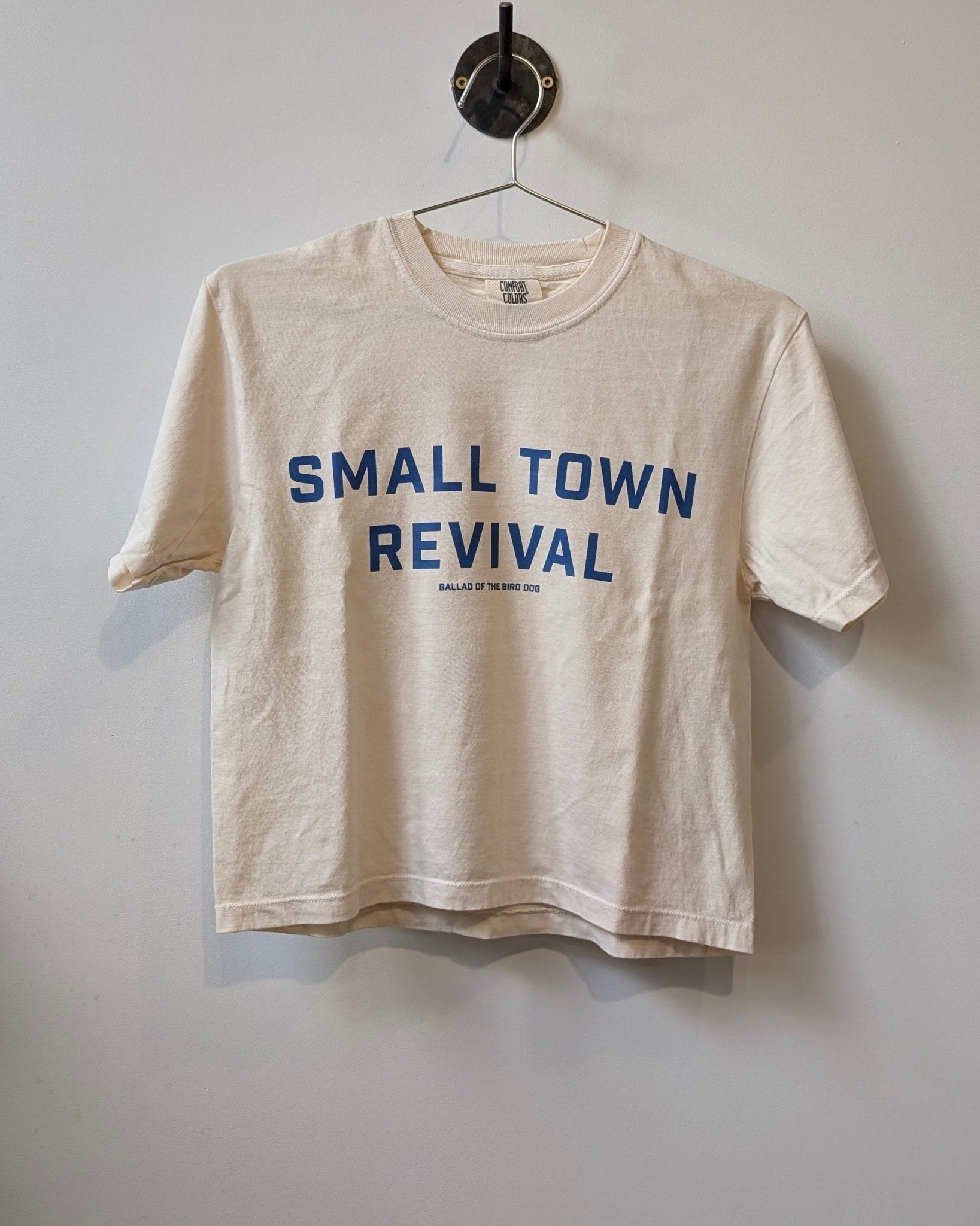 A beige Shop Shirt | Small Town Revival Crop by Ballad of the Bird Dog hangs on a hanger, with bold blue text celebrating small town community and smaller text beneath.