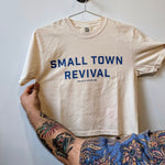 A tattooed person holds the Ballad of the Bird Dog "Shop Shirt | Small Town Revival Crop," a handcrafted cotton shirt with "SMALL TOWN REVIVAL" in blue letters, displayed on a hanger against a plain background.
