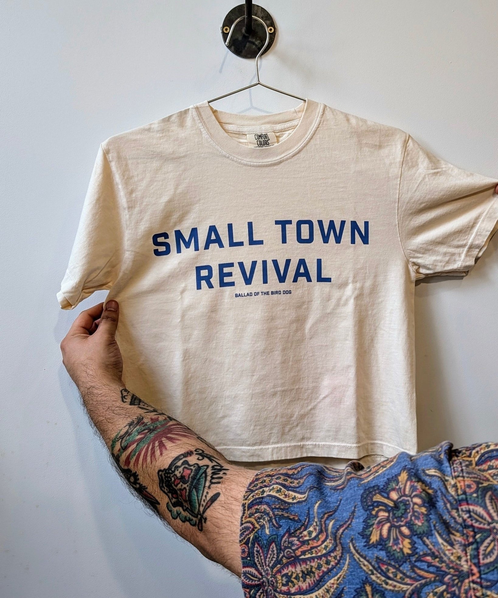 A tattooed person holds a beige Ballad of the Bird Dog "Shop Shirt | Small Town Revival Crop," featuring blue "SMALL TOWN REVIVAL" text—a tribute to small town community.