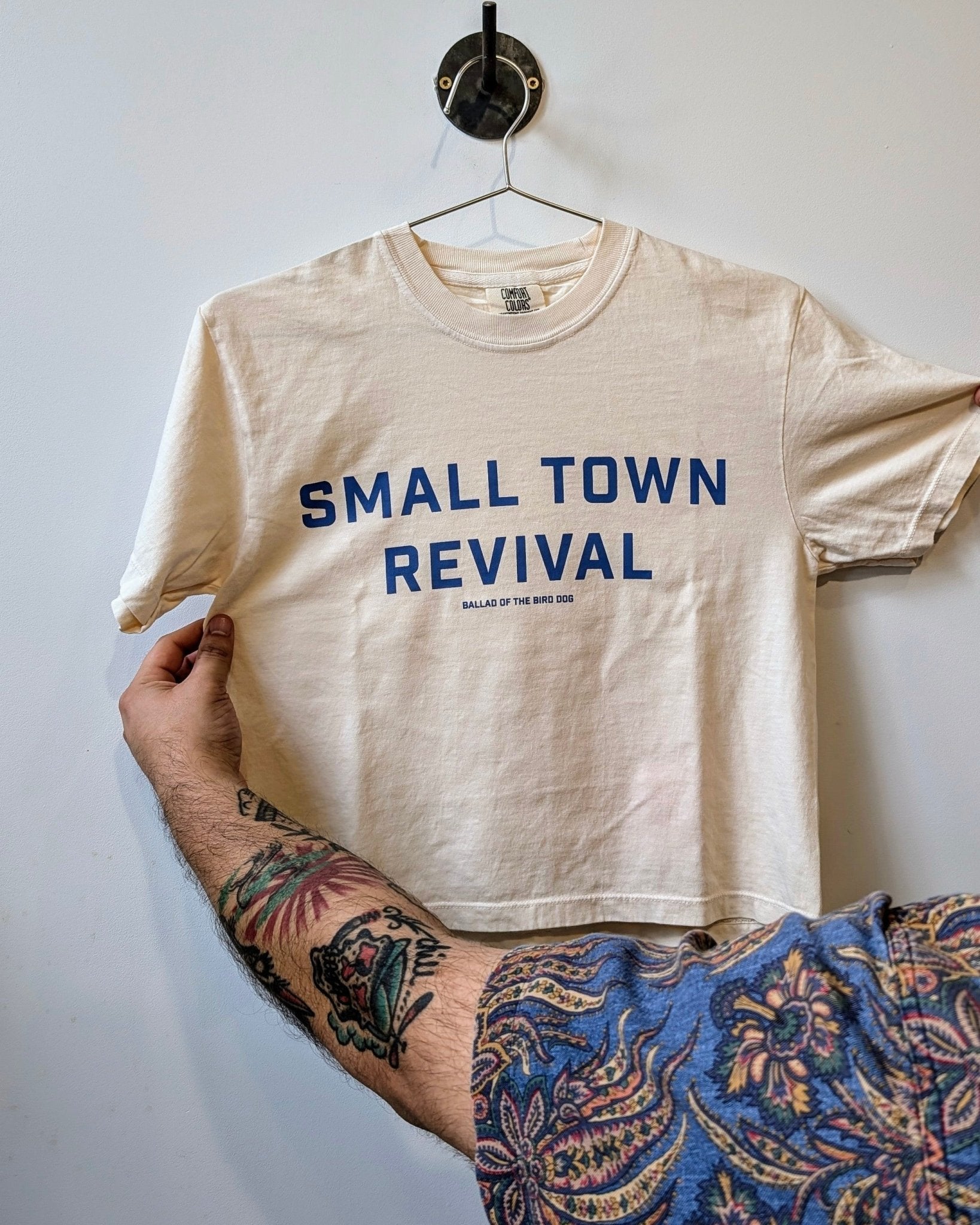 A tattooed person holds a beige Ballad of the Bird Dog "Shop Shirt | Small Town Revival Crop," featuring blue "SMALL TOWN REVIVAL" text—a tribute to small town community.