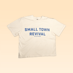 The Shop Shirt | Small Town Revival Crop by Ballad of the Bird Dog is a handcrafted white cotton shirt featuring “SMALL TOWN REVIVAL” in bold blue letters, celebrating community spirit on a beige background.