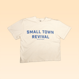 The Ballad of the Bird Dog Shop Shirt | Small Town Revival Crop features bold blue "SMALL TOWN REVIVAL" text on white, laid flat on beige—a perfect crop shirt for showing off your small town pride.