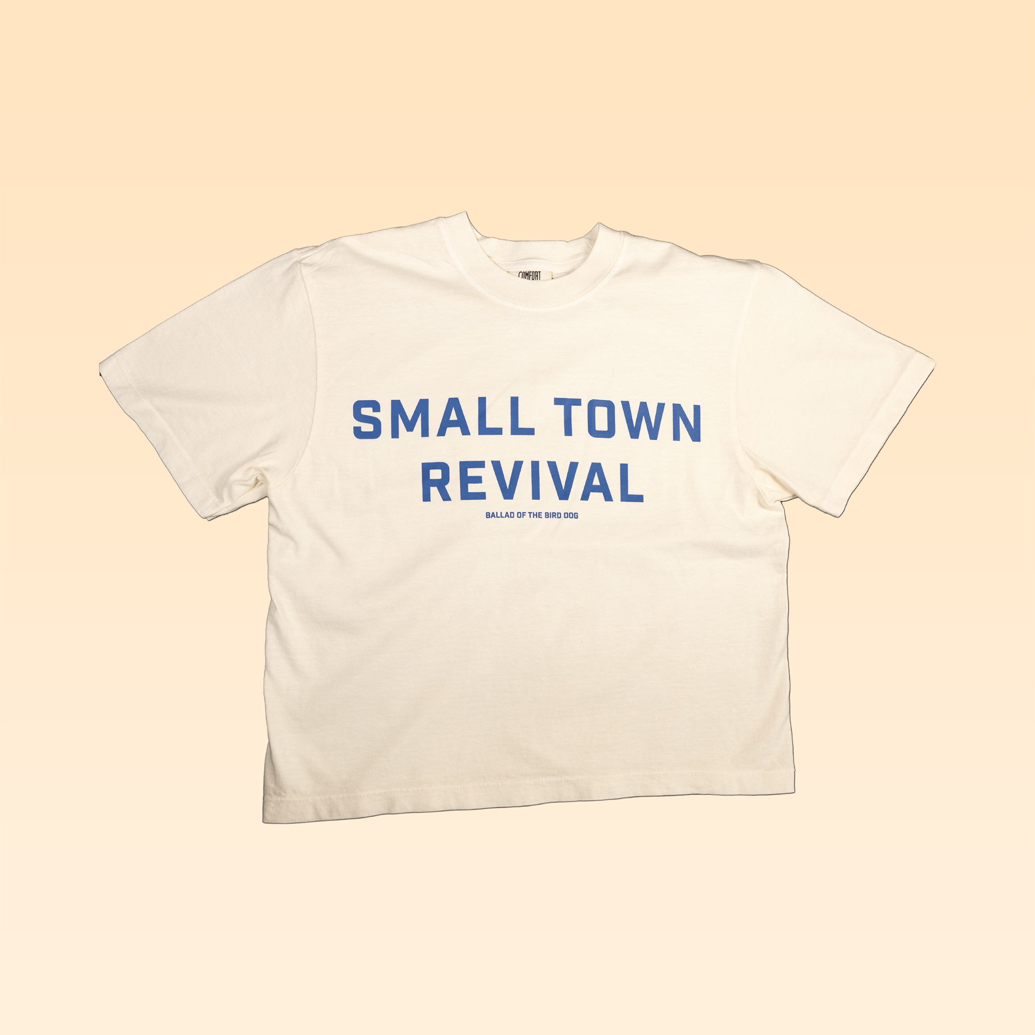 The Ballad of the Bird Dog Shop Shirt | Small Town Revival Crop features bold blue "SMALL TOWN REVIVAL" text on white, laid flat on beige—a perfect crop shirt for showing off your small town pride.