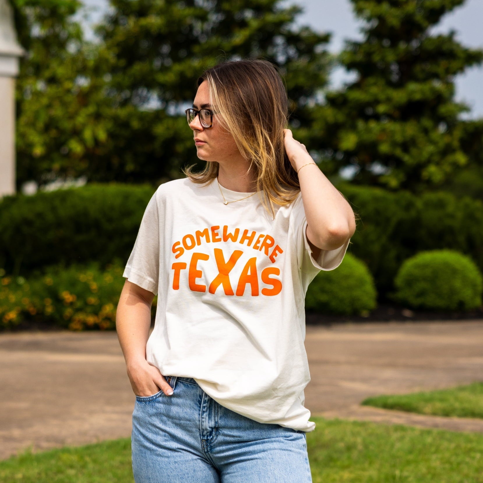 A person stands outdoors in blue jeans and a white Ballad of the Bird Dog Shop Shirt | Somewhere Texas, embodying small-town Texas charm. Lush green trees and bushes provide a picturesque backdrop.