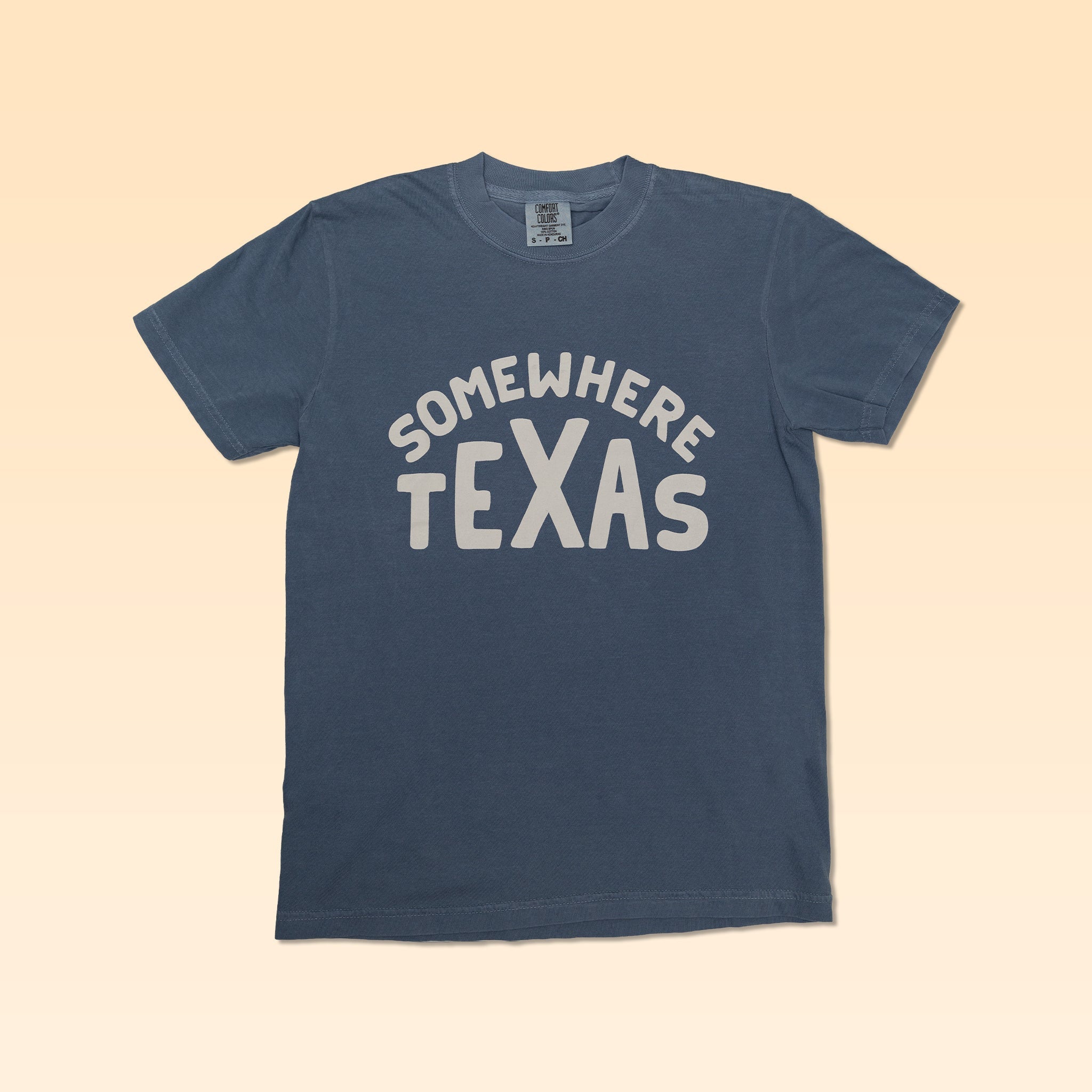 The Shop Shirt | Somewhere Texas by Ballad of the Bird Dog is a blue short-sleeve tee, ideal for fans of small-town Texas style, shown on a light beige gradient background.