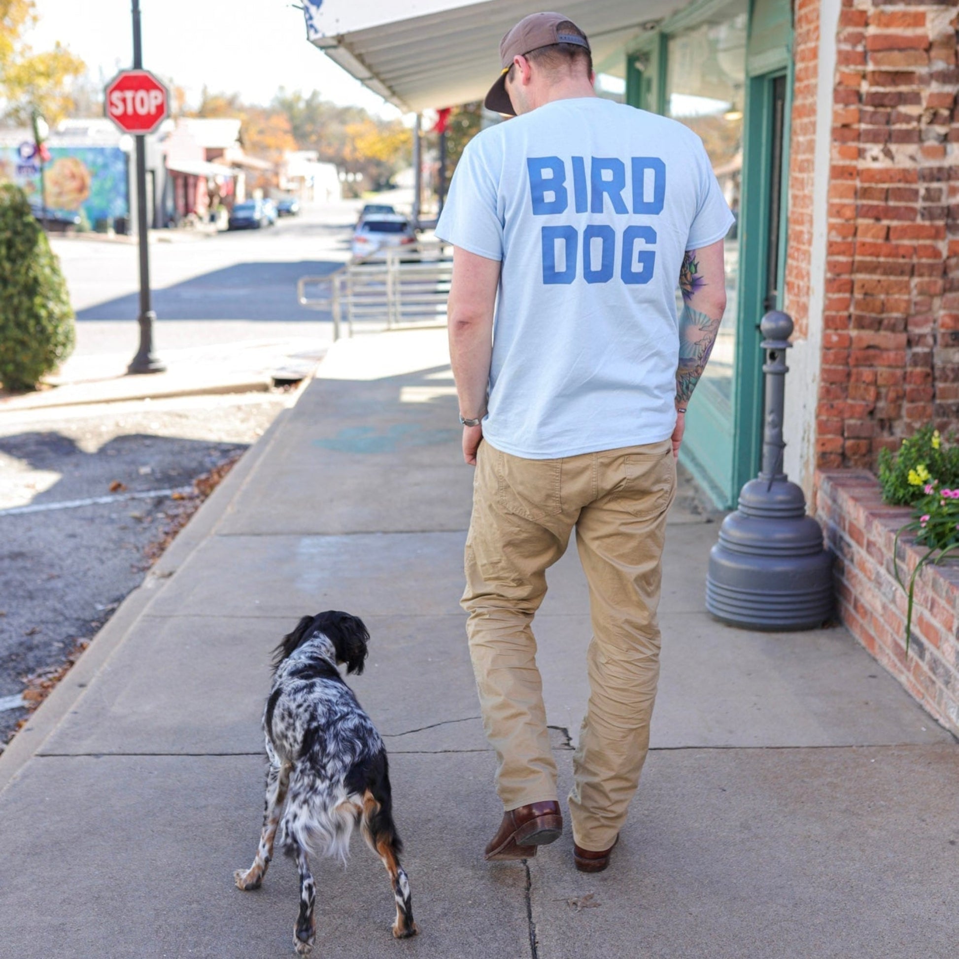 A man in the "Shop Shirt | Team Bird Dog" by Ballad of the Bird Dog walks a black and white dog on a small town sidewalk.