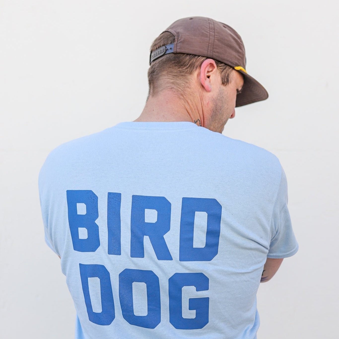 A man wears a brown cap and the Ballad of the Bird Dog "Shop Shirt | Team Bird Dog," a light blue baseball-style jersey with "BIRD DOG" boldly on the back.