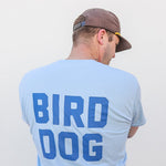 Someone wearing the light blue "Team Bird Dog" Shop Shirt by Ballad of the Bird Dog, featuring bold "BIRD DOG" lettering on the back, stands against a plain white background.