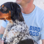 A person in a light blue "Shop Shirt | Team Bird Dog" from Ballad of the Bird Dog sits beside a black and white dog with brown markings.