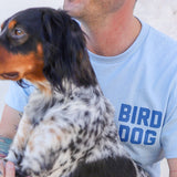Wearing the light blue Shop Shirt by Ballad of the Bird Dog, a person sits with a black and white dog with brown markings, proudly showing their love for Team Bird Dog apparel.