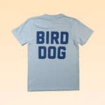 The Shop Shirt by Ballad of the Bird Dog features bold blue "BIRD DOG" text on the back. Laid flat on beige, this Team Bird Dog shirt is a sporty pick for any bird dog fan's collection.