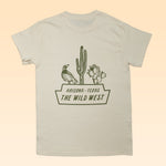 The Shop Shirt | The Wild West by Ballad of the Bird Dog is a beige tee with a green bird, cacti, and "ARIZONA - TEXAS THE WILD WEST" graphic on the back—perfect for desert wildlife fans.