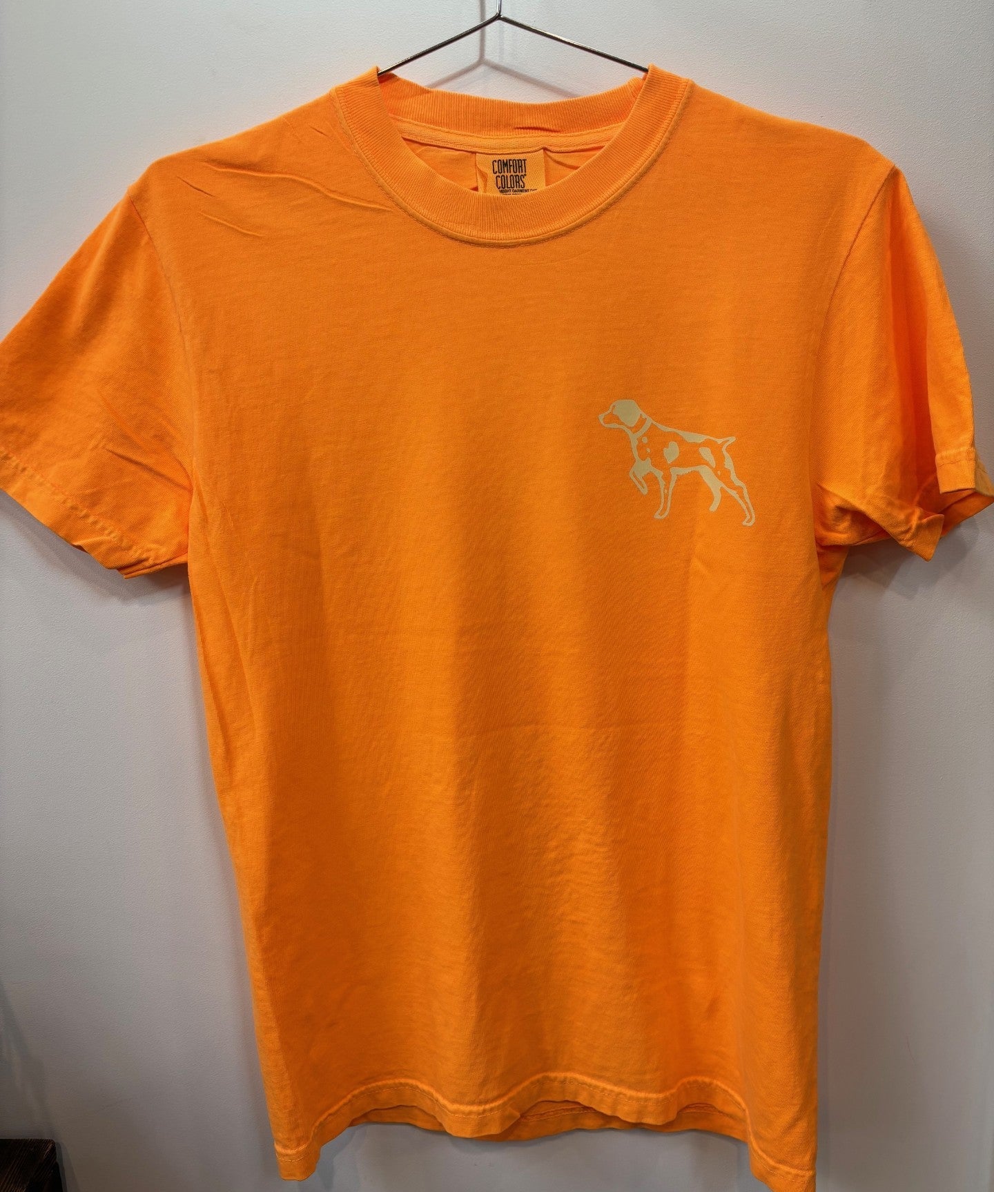 The Ballad of the Bird Dog Shop Shirt | Upland is an orange short-sleeve tee featuring a small outlined dog graphic on the left chest, shown on a hanger against a white wall.