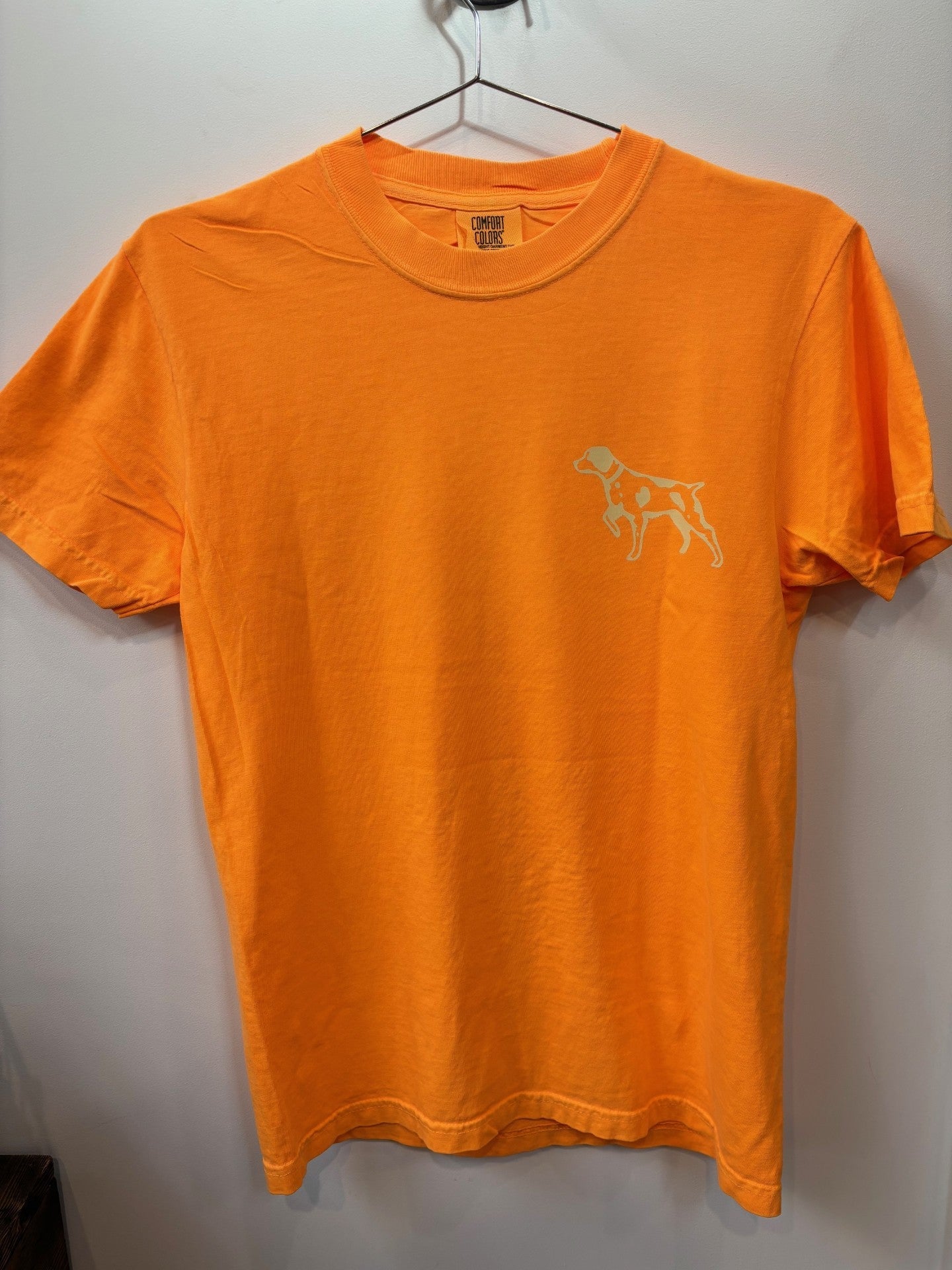 The Ballad of the Bird Dog Shop Shirt | Upland is an orange short-sleeve tee featuring a small outlined dog graphic on the left chest, shown on a hanger against a white wall.