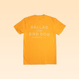 The Ballad of the Bird Dog "Shop Shirt | Upland" is a yellow tee with “BALLAD of the BIRD DOG BRENHAM, TX” in white on the back, shown on a light peach background.