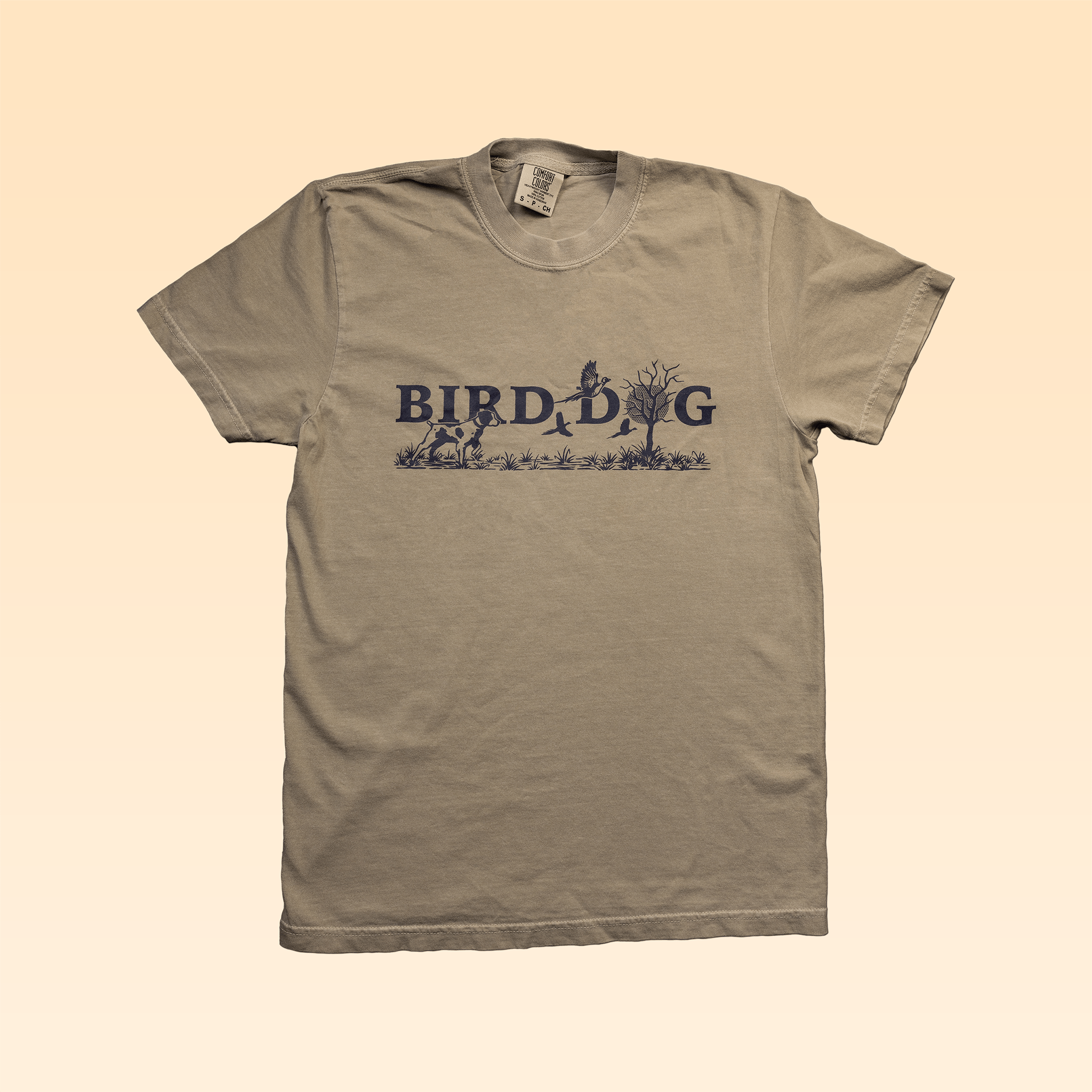 The Ballad of the Bird Dog Shop Shirt in Upland Morning is a beige, USA cotton tee, garment-dyed for comfort and features “BIRD DOG” with a dark blue dog, tree, and bird graphic across the chest.
