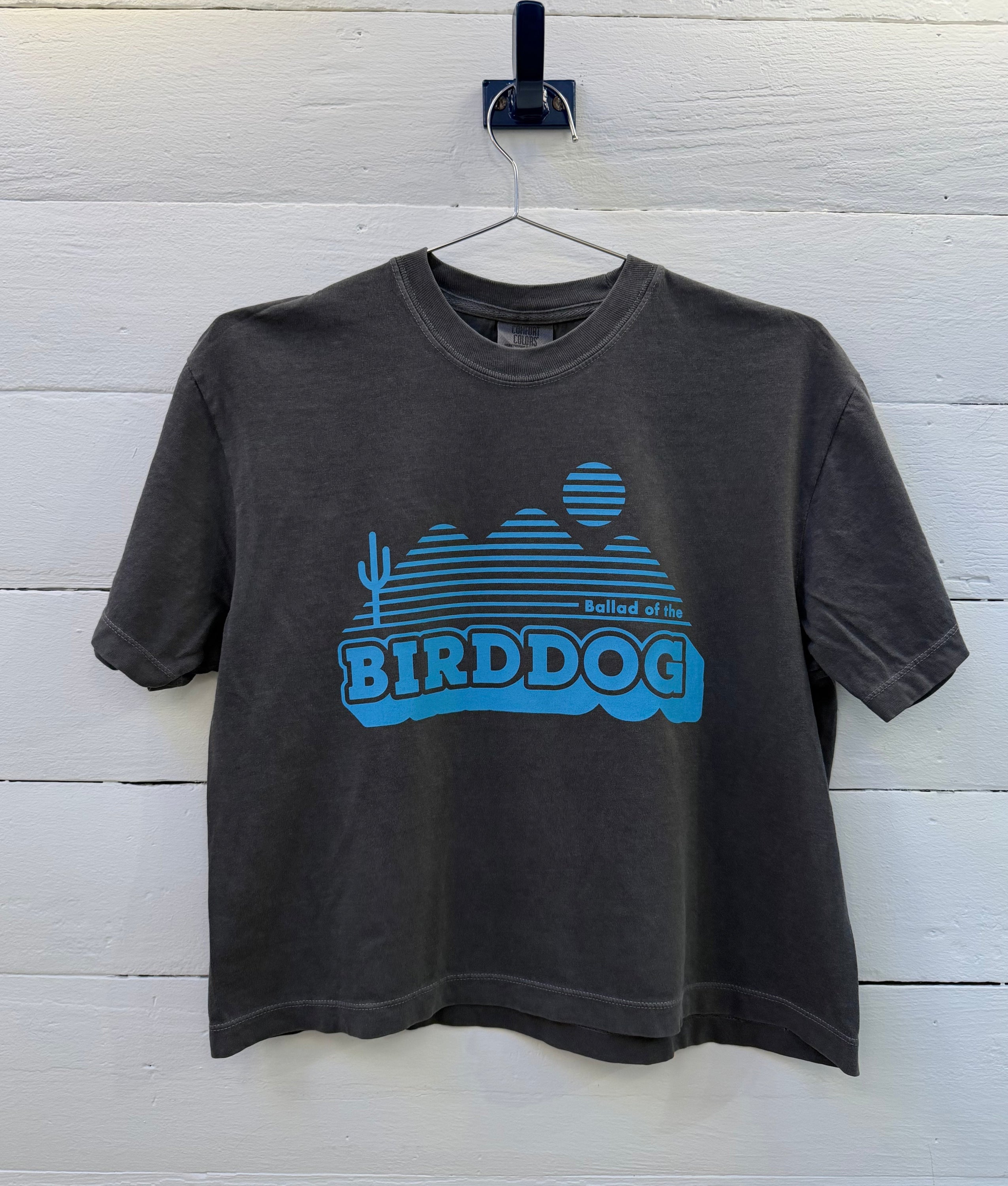 The Ballad of the Bird Dog Shop Shirt | Vintage Bird Dog Crop is a dark gray tee on a hanger, showing a blue mountain, cactus, and setting sun graphic with "Birddog" text on the front.
