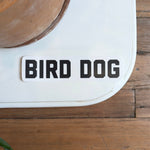 The Shop Sticker | Bird Dog Honor by Ballad of the Bird Dog features bold black "BIRD DOG" text on a rectangular sticker—ideal for hunting and outdoor adventure fans. Shown on a white surface beside a brown plant pot on a wooden floor.