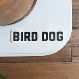 The Shop Sticker | Bird Dog Honor by Ballad of the Bird Dog features bold black "BIRD DOG" text on a rectangular sticker—ideal for hunting and outdoor adventure fans. Shown on a white surface beside a brown plant pot on a wooden floor.