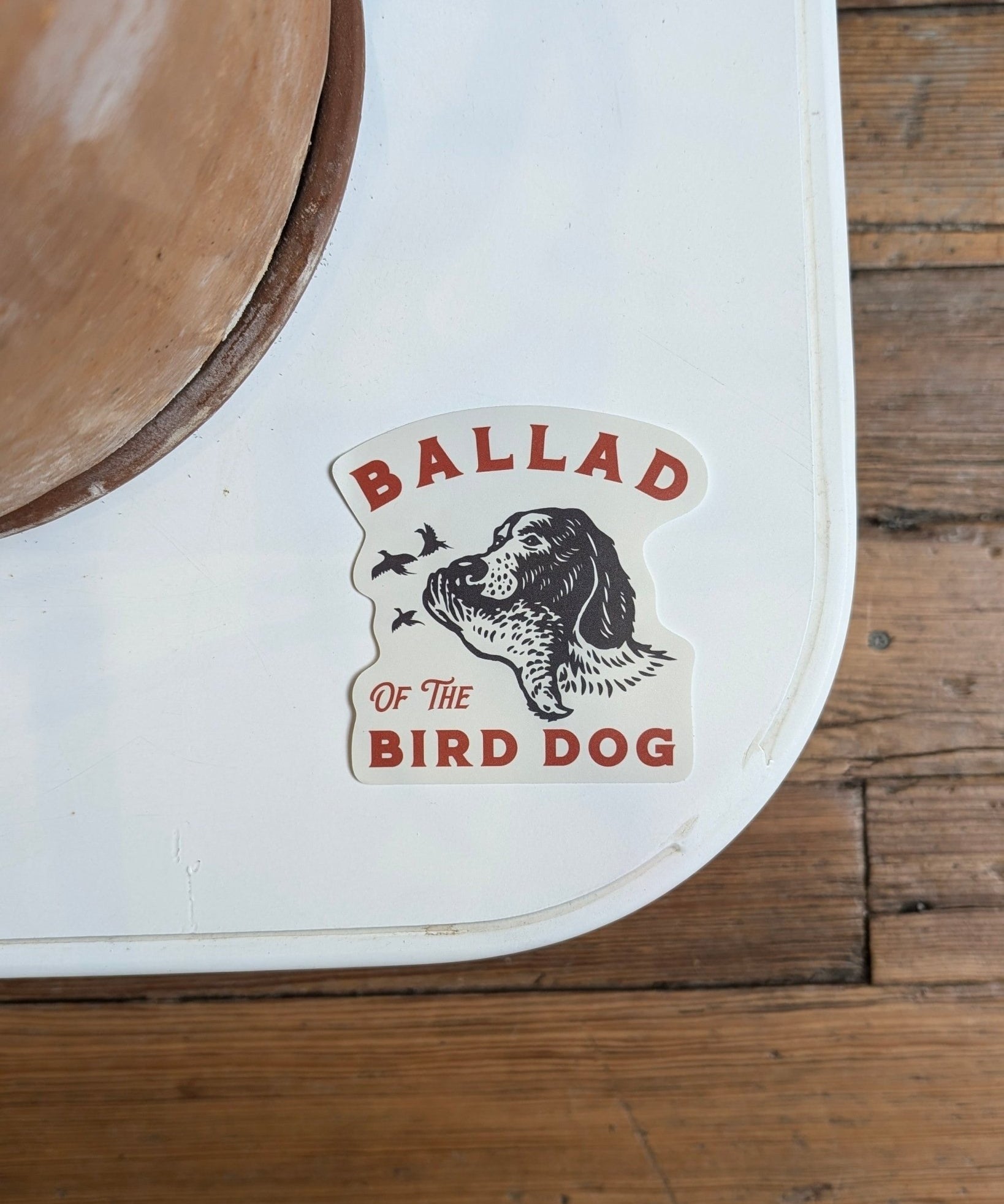 The Shop Sticker | Retriever by Ballad of the Bird Dog features a dog watching birds with "Ballad of the Bird Dog" in red and black on a white background over wooden floorboards.