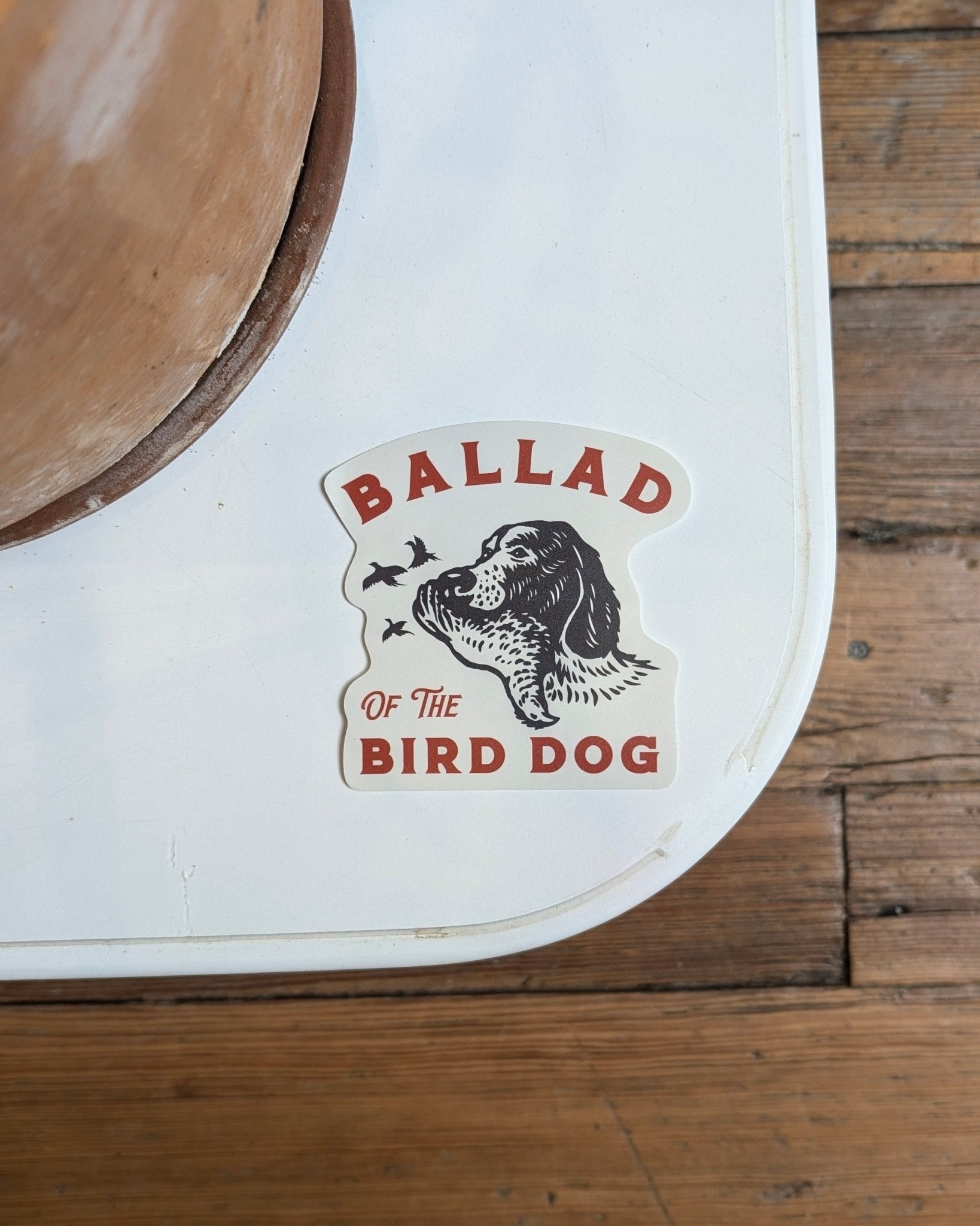 The Shop Sticker | Retriever by Ballad of the Bird Dog features a dog watching birds with "Ballad of the Bird Dog" in red and black on a white background over wooden floorboards.
