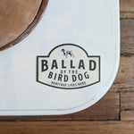 A white surface displays the Ballad of the Bird Dog "Heritage Lives Here" Shop Sticker with a dog silhouette; part of a wooden floor is visible.