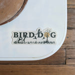 The Shop Sticker | Upland Morning by Ballad of the Bird Dog features "BIRD DOG" spelled out with dog, birds, plants, and tree illustrations—celebrating upland hunting traditions—seen here on a white surface near a wood floor.
