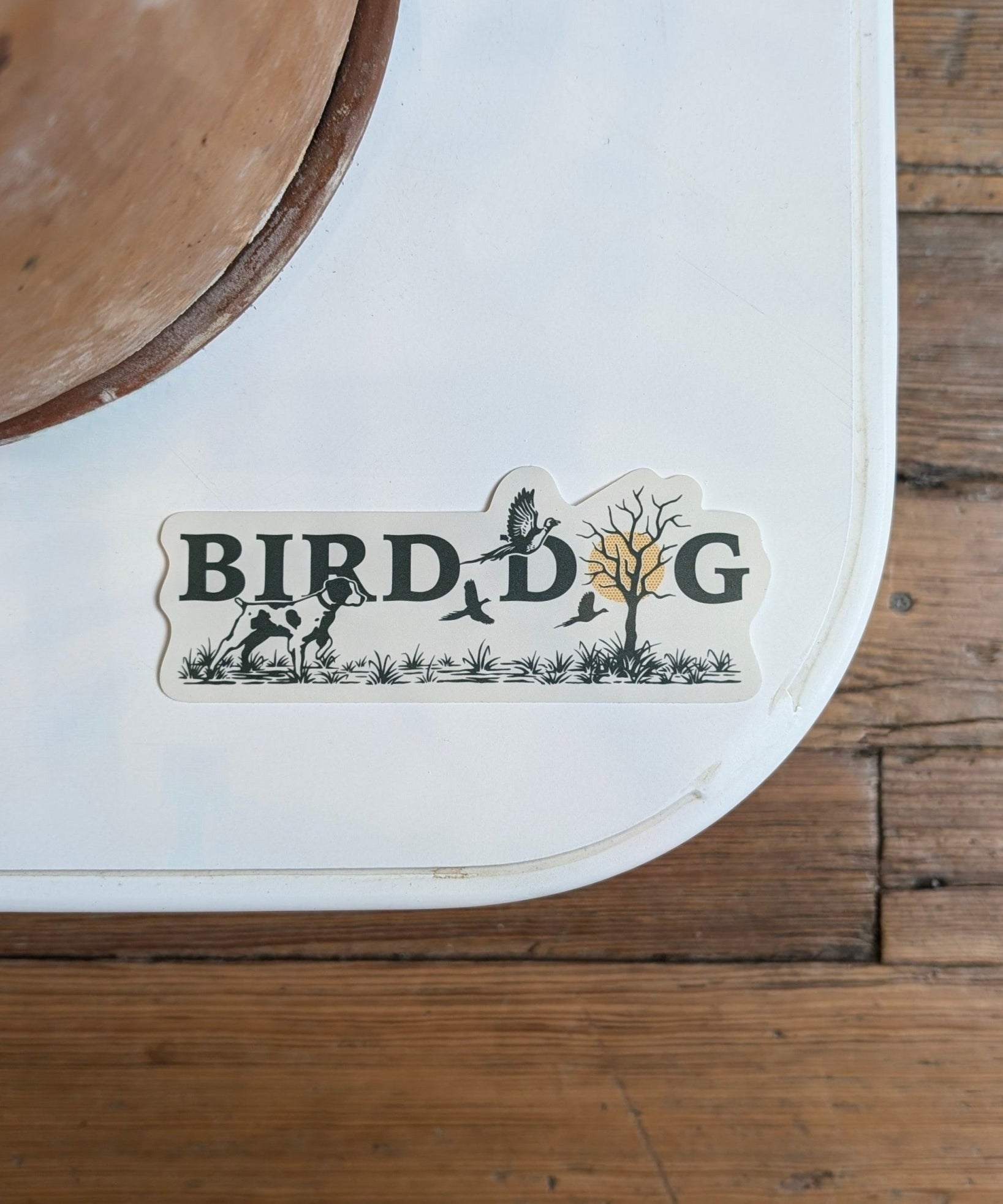 The Shop Sticker | Upland Morning by Ballad of the Bird Dog features "BIRD DOG" spelled out with dog, birds, plants, and tree illustrations—celebrating upland hunting traditions—seen here on a white surface near a wood floor.