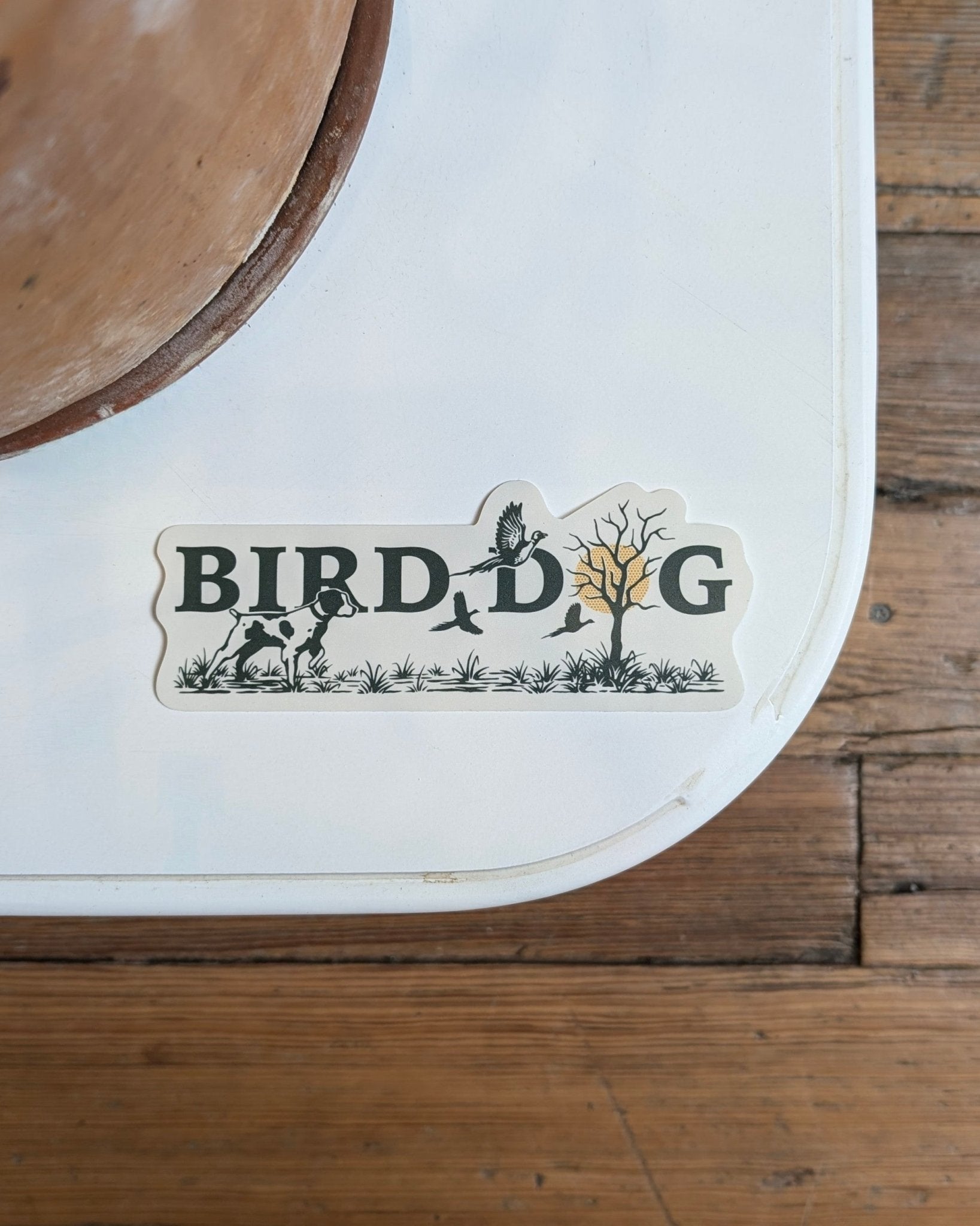 The Shop Sticker | Upland Morning by Ballad of the Bird Dog features "BIRD DOG" spelled out with dog, birds, plants, and tree illustrations—celebrating upland hunting traditions—seen here on a white surface near a wood floor.